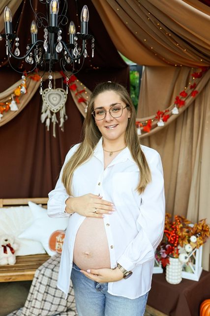 Uśmiechnięta kobieta w ciąży w białej koszuli trzyma dłonie na brzuszku