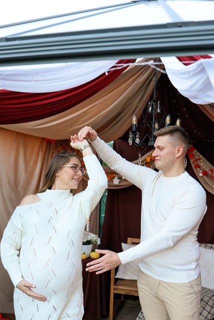 Mężczyzna obraca w tańcu ciężarną partnerkę w białym swetrze