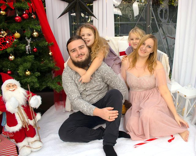 Tata z córką na plecach i mama z drugą córką na białym kocu przy choince