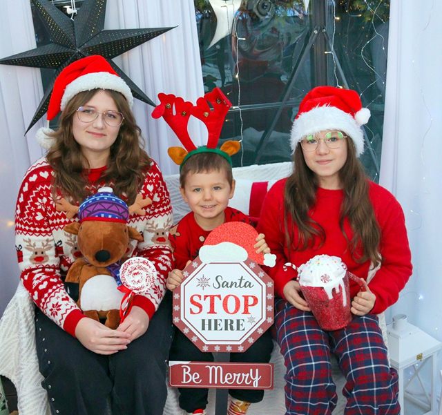 Troje dzieci w mikołajowych czapkach z tabliczką Santa Stop Here
