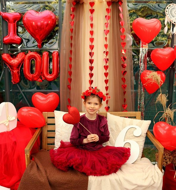 Dziewczynka w bordowej tiulowej sukience na ławce, w tle balony „I love you”