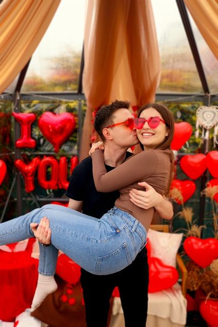 Mężczyzna trzyma partnerkę na rękach wśród czerwonych serc i balonów „I love you”