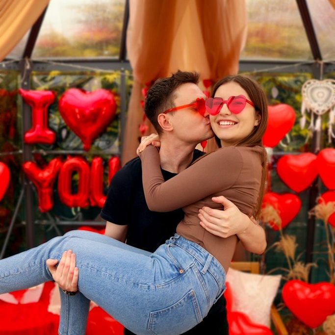 Mężczyzna trzyma partnerkę na rękach wśród czerwonych serc i balonów „I love you”